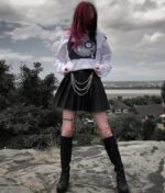 A person with red-tinted hair stands on a rocky surface, wearing a HellStar Discover Sweater, pleated skirt with chain accessories, fishnet stockings, and boots. The background features trees, houses, and a cloudy sky.